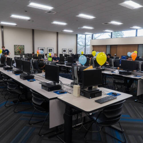 A computer lab with rows of desktop computers on long tables, each station decorated with colorful balloons. Several people are seated at the computers, working or talking, while a few individuals are standing or walking around.